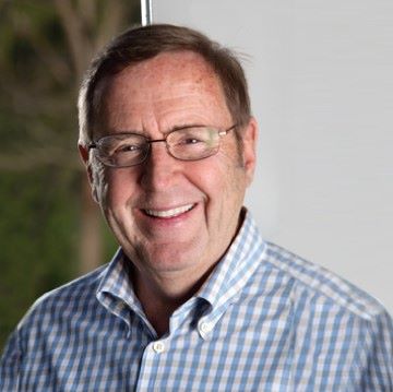 David Gaw, a white man with short brown hair and glasses, smiles at the camera wearing a white and blue checkered dress shirt.