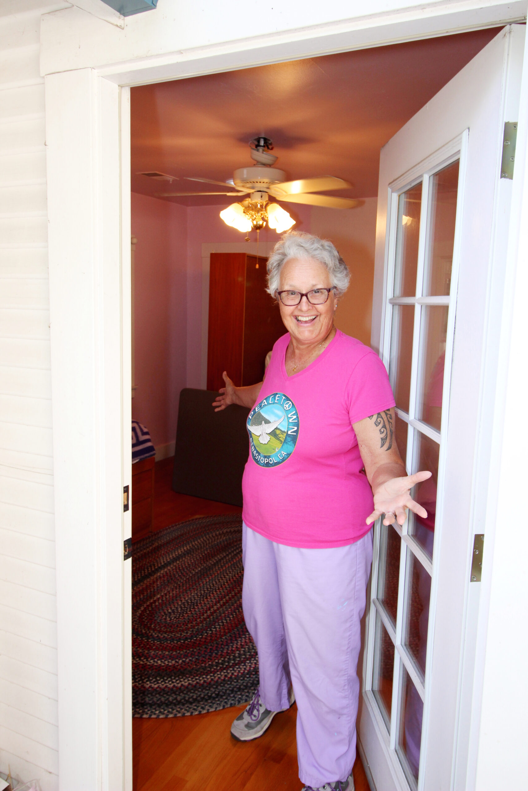 A woman with short white curly hair smiles in her ADU, wearing a pink t-shirt and purple pants.