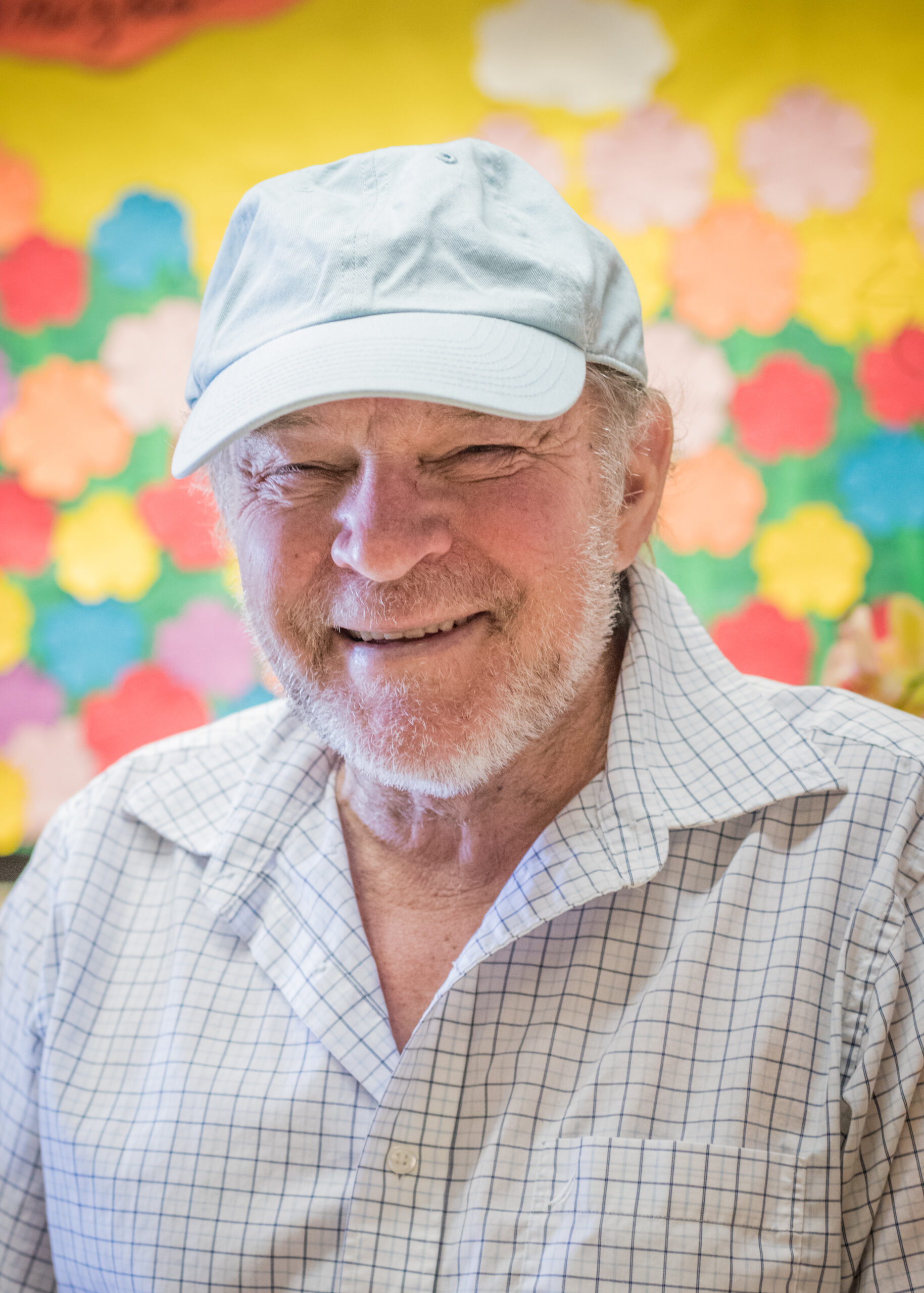 An image of an older man with short grey hair and facial hair, wearing a hat and a checkered collared shirt, smiling.