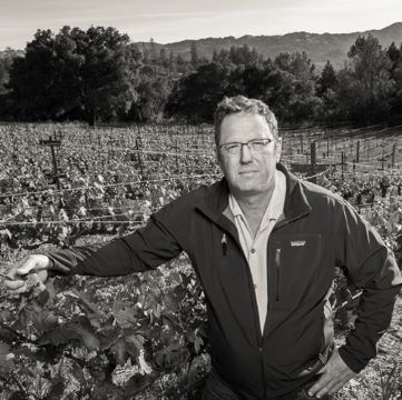 A black and white image of Pete Richmond, a white man with short brown hair, stands in a vineyard, wearing a collared shirt under a Patagonia jacket