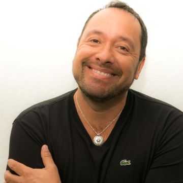 Oscar Renteria, a Latino man with short dark hair and facial hair, smiles at the camera wearing a silver necklace and a black t-shirt