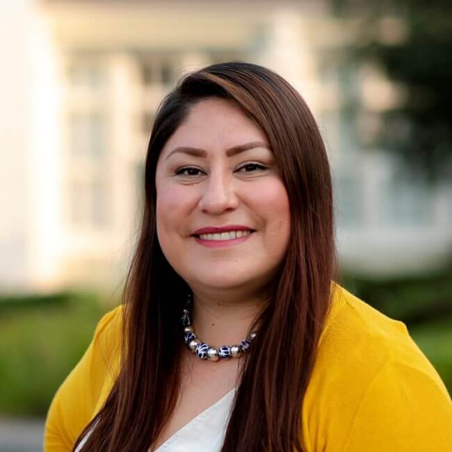 Elba Gonzalez-Mares, a Latina woman smiles at the camera wearing a white blouse under a yellow cardigan, wearing a blue, white, and silver necklace.