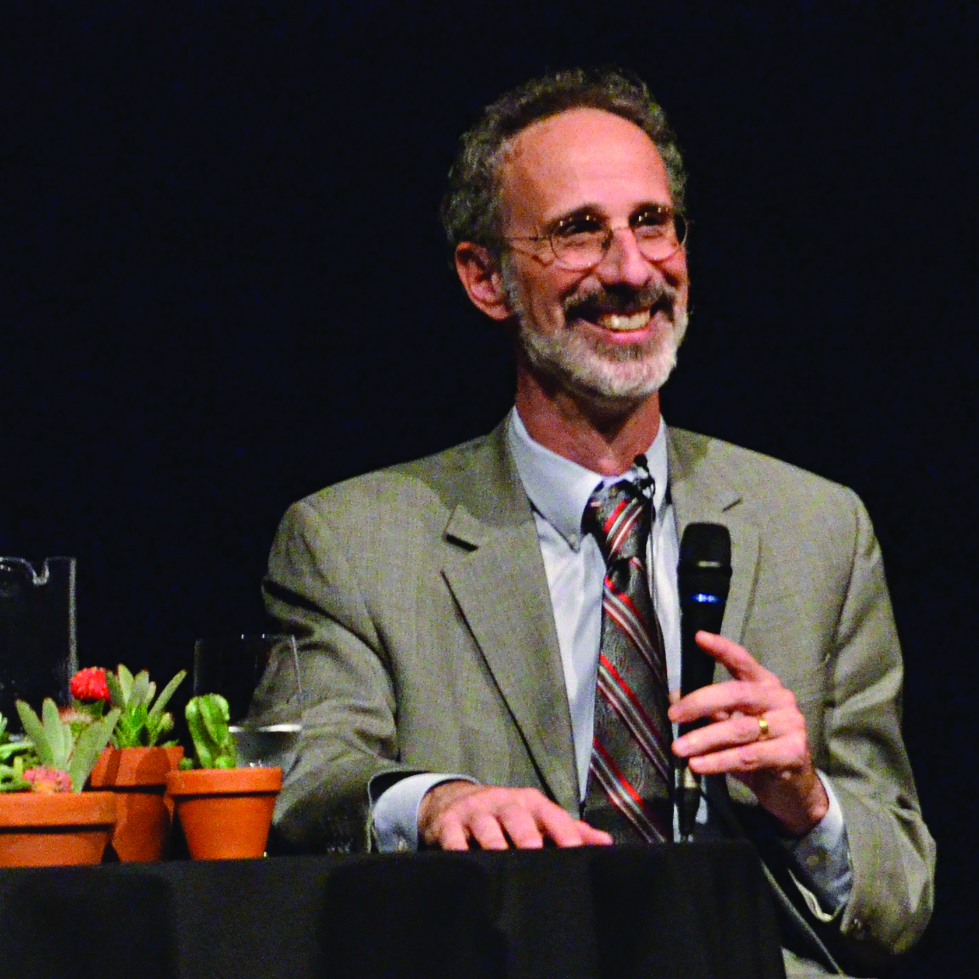 A photo of Dr. Peter Gleick, a middle aged white man smiles looking away from the camera, holding a microphone wearing glasses and a suit and tie