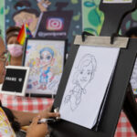 A portrait artist draws a caricature of a young person at a booth at the pride festival