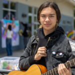 A young person with poses with a guitar and two pride pins on their jacket.