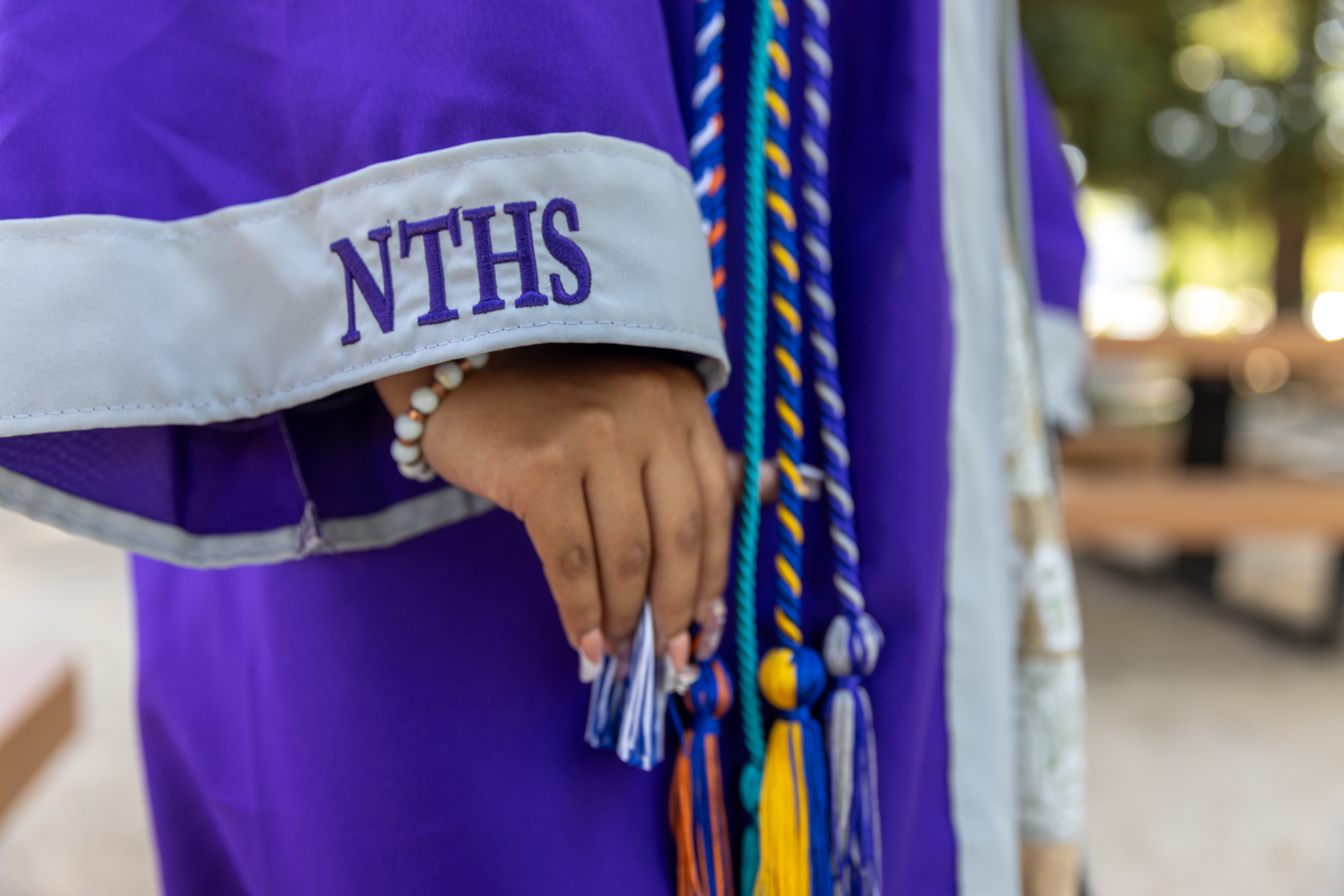 A closeup photo of Araceli's graduation gown. The sleeve has embroidery reading NTHS, and she has a number of colorful tassels around her neck.