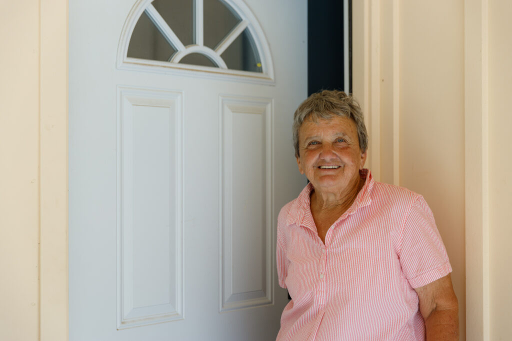 A person with short hair and a pink shirt, representing the Napa Valley Community Foundation (NVCF), smiles while standing next to an open white front door with decorative windows at the top. The light-colored wall in the background adds to the welcoming scene.
