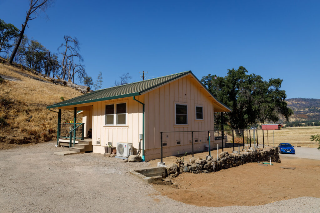 A small beige house with a green roof sits on a dry, gravel lot embraced by the rustic charm of Napa Valley. A blue car rests nearby, while a stone-lined path meanders to the entrance. The clear sky mirrors the community spirit fostered by NVCF amidst the brown hills and sparse trees.