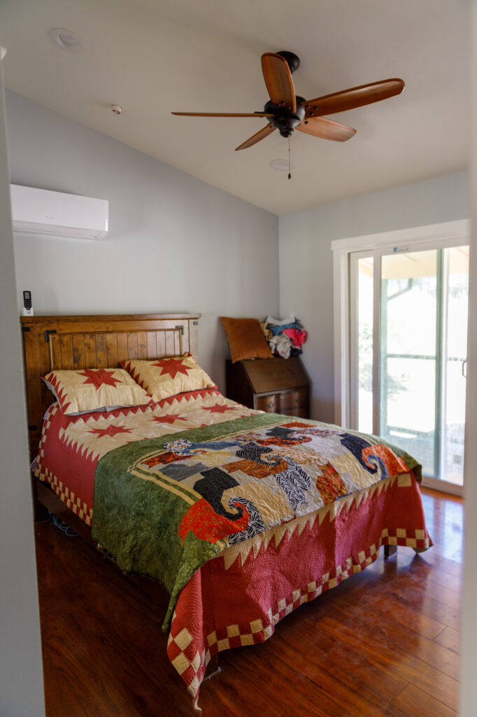 A cozy bedroom with a wooden bed draped in a colorful quilt featuring various patterns. A ceiling fan whirrs above, while a small wooden desk sits in the corner. The sliding glass door lets NVCF's natural light flood in, illuminating the hardwood flooring beneath.