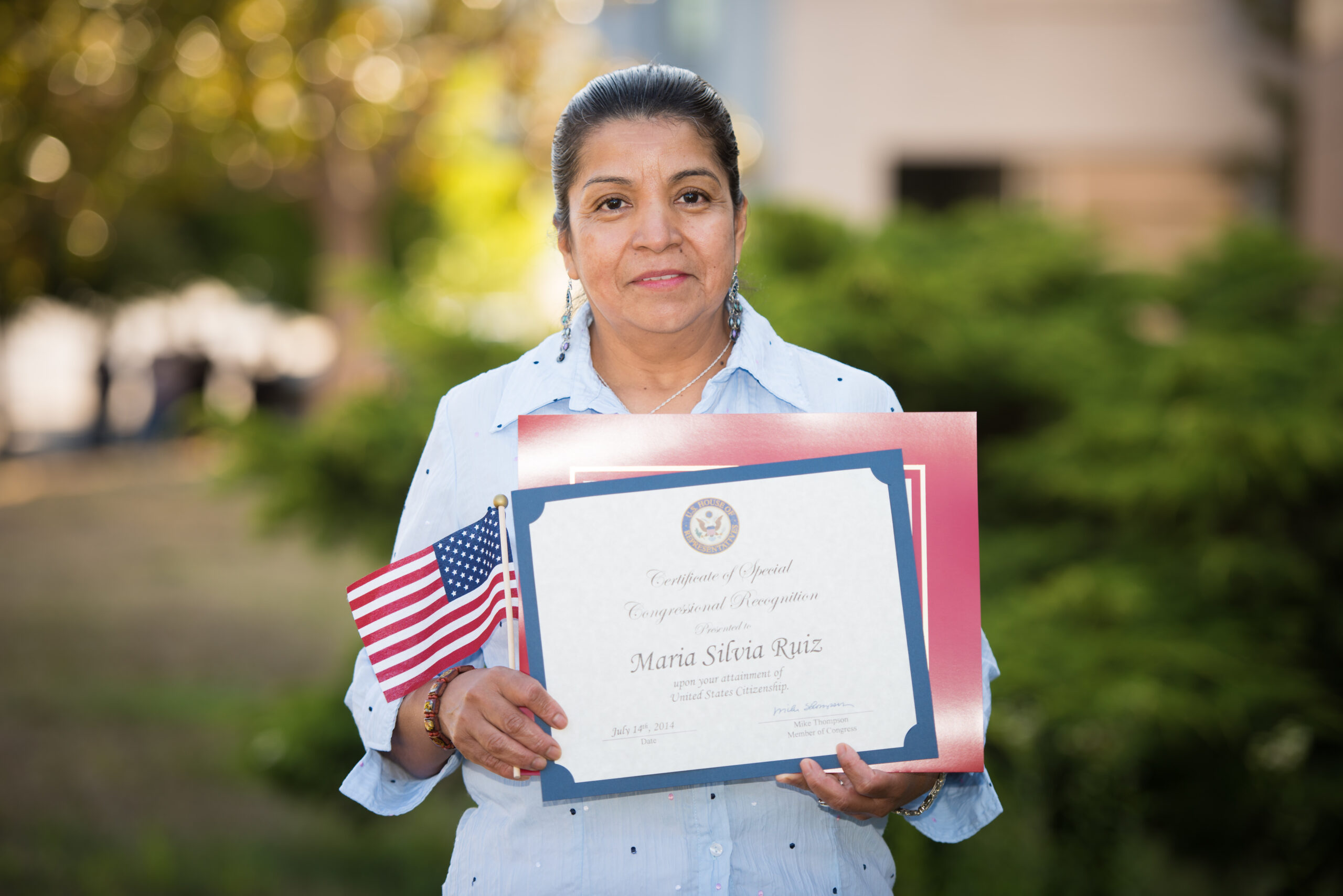 A woman, proudly supported by the Napa Valley Community Foundation, holds a certificate and a small American flag outdoors. Wearing a light blue shirt, she stands amidst lush greenery, with buildings artistically blurred in the background.