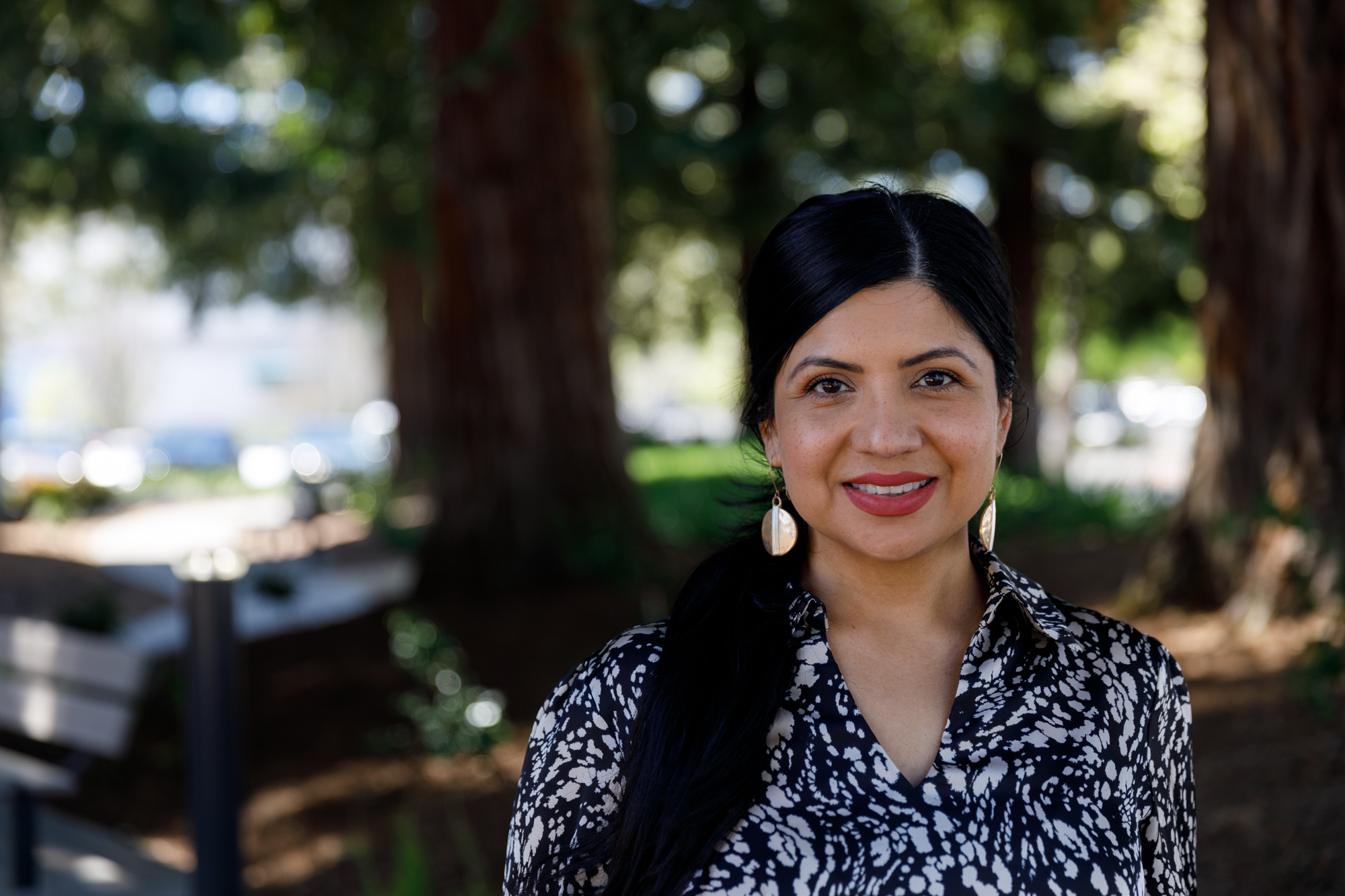 Maria Pantoja is a Latina woman with long brown hair and a beautiful, welcoming smile. She is standing outdoors, and is wearing a chic black and white top with gold earrings.