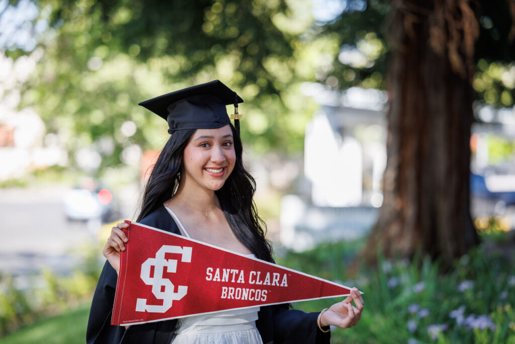 Arisbeth Lopez is wearing her block colored graduation cap and gown, and a white dress. She is a young Latina woman, and has a big smile on her face, and is holding a banner reading "Santa Clara Broncos". Arisbeth is a graduate of American Canyon High School and recipient of the Daisy Cavalli Scholarship, is enrolled at Santa Clara University, where she will study International Business.