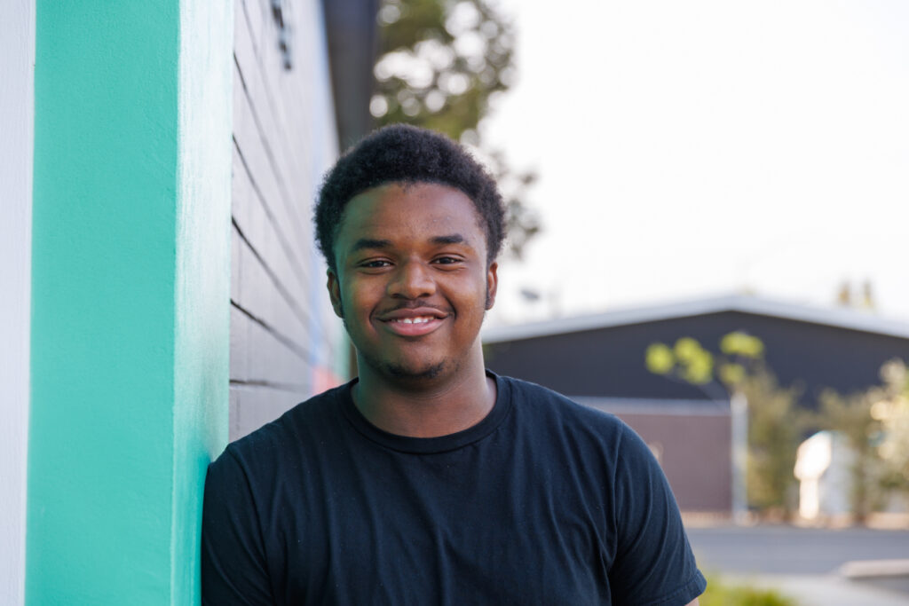 Evan Smith is a young man with a big smile. He is Black, and is posing in front of Napa Valley Community Foundation's office wearing a black colored t-shirt. Evan is attending Solano Community College’s Automotive Technology program with support from NVCF’s Skilled Trades Scholarship. He hopes to one day open his own shop.