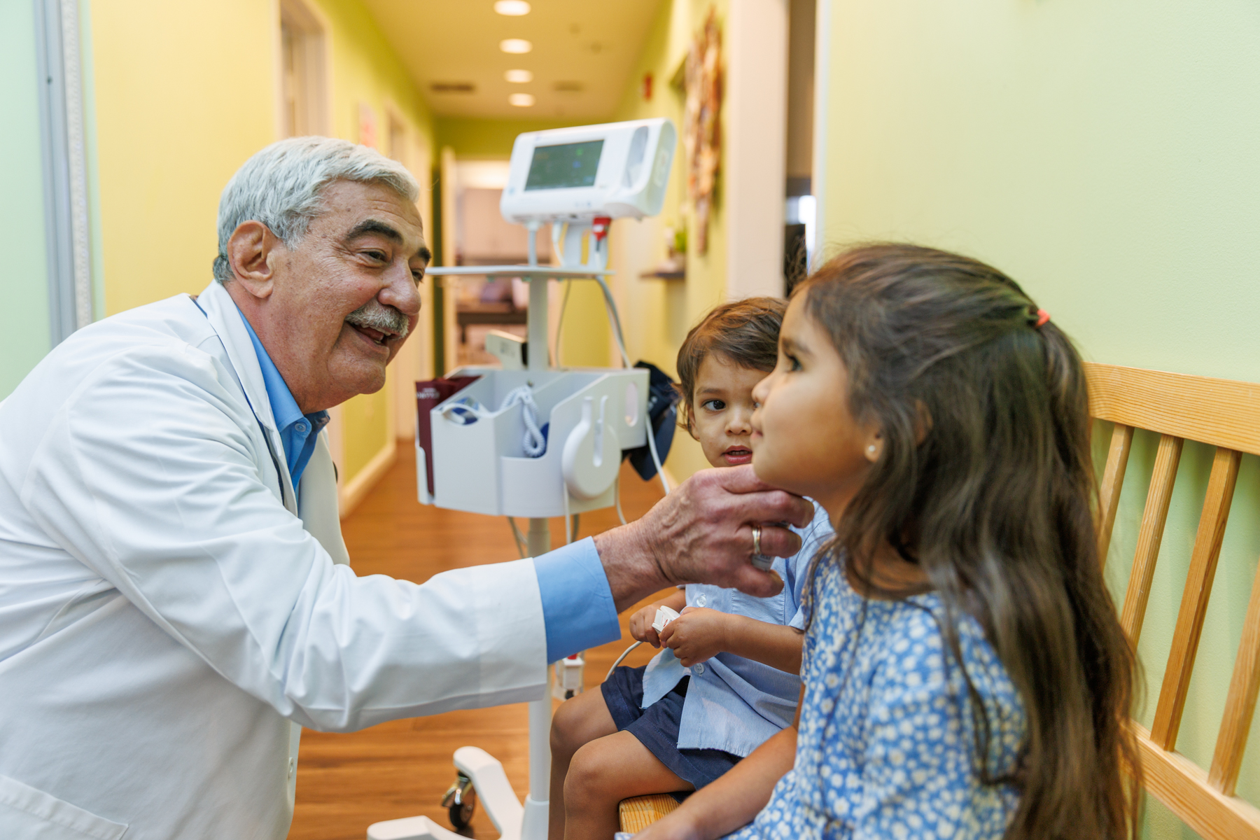 Doctor Joseph Carrillo examines a young girl’ while her brother sits beside her on a wooden bench in a medical clinic hallway. Medical equipment is visible in the background.